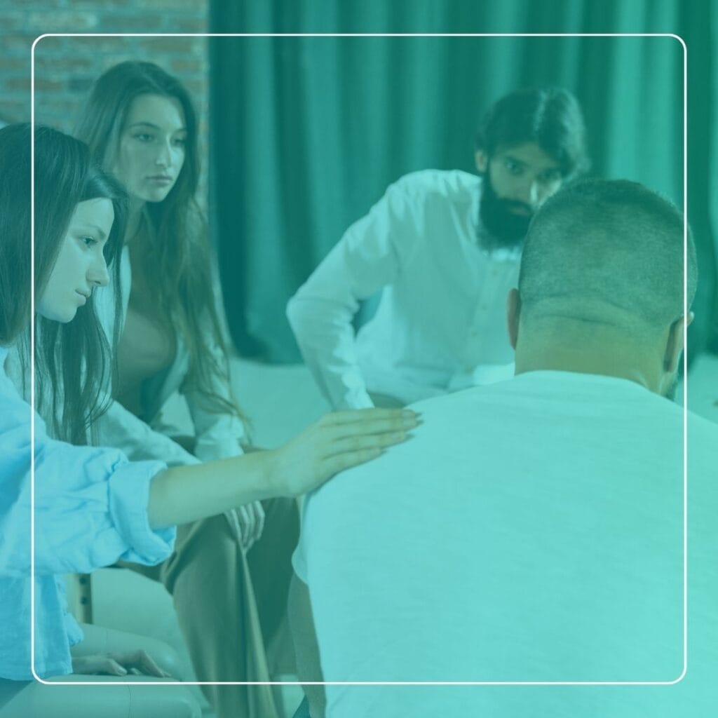 This photograph captures a small group of people seated in a circle during a group therapy session, with one participant placing a hand on another's shoulder in a gesture of support. Such sessions are often used to foster peer connection, encourage open dialogue, and provide emotional and practical assistance to members.